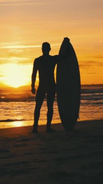 Gün batımında sörf tahtasıyla sahilde duran formda bir adamın silüeti. Orta çekim