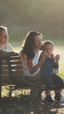 Aile bir bankta oturuyor ve küçük bir çocuk sabun köpüğüyle oynuyor. Orta çekim