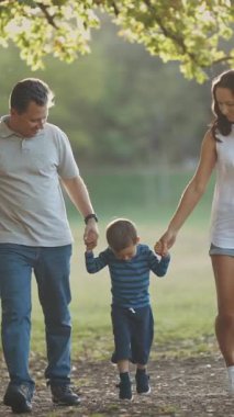 Parkta yürüyen sevimli genç bir aile. Ebeveynler küçük oğullarını ellerinden tutuyorlar. Orta çekim