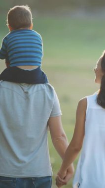 Parkta yürüyen sevimli genç bir aile. Babasının omuzlarında küçük bir çocuk. Orta çekim