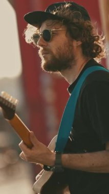 Duvarın yanında durup gitar çalan şık bir genç adam. Orta çekim