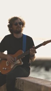Rıhtımda oturan ve gün batımında gitar çalan bir adam. Orta çekim