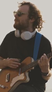 Gün batımında limanda gitar çalan şık bir genç adam. Orta çekim