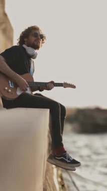 Rıhtımda oturan ve gün batımında gitar çalan bir adam. Orta çekim