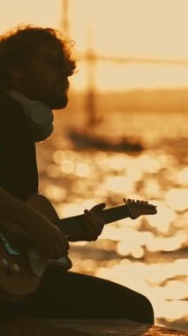 Kablosuz kulaklık takan bir adam gün batımında rıhtımda oturup gitar çalıyor. Orta çekim