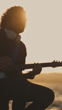 Sahilde gün batımında gitar çalan yalnız bir genç adam. Orta çekim