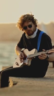 Genç, yalnız bir gitarist, bacaklarını suya sarkıtarak şehir sehpasında çalıyor. Orta çekim