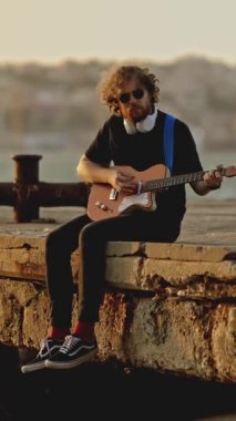 Bir adam gün batımında sette bacakları suya sarkık oturur ve gitar çalar. Orta çekim