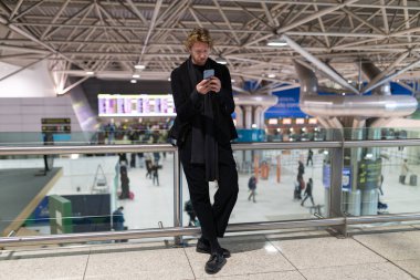 Modern havaalanı terminalinde beklerken akıllı telefon kullanan genç bir adam.