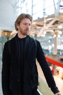 Sakallı ve kıvırcık saçlı bir adam modern bir binada tefekkür ediyor.