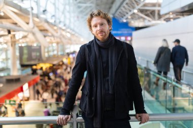 Modern bir havaalanı terminalinde duran genç adam, arka planda gezginler.