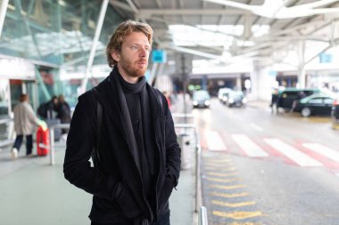 Genç adam havaalanında dikiliyor, beklerken düşünceli görünüyor.