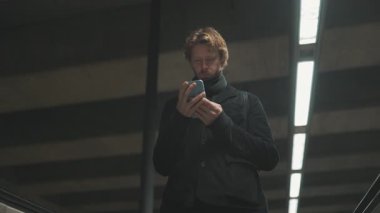 Siyah ceketli genç adam metroda yürüyen merdivenden çıkarken telefonuna bakıyor.