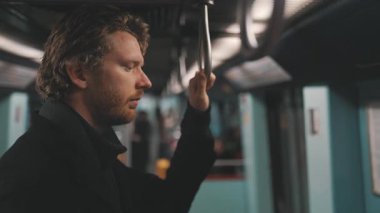 Düşünceli genç bir adam, gözleri kapalı metro vagonunda duruyor, tırabzana tutunuyor.