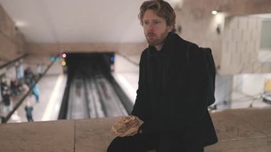 Modern bir metro istasyonunda treni beklerken sırt çantalı bir adam sandviç yiyor.