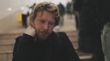 Arka planda bir grup insan geçerken üzgün, düşünceli genç adam merdivenlerde oturuyor.