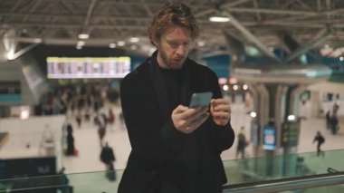 Stresli genç adam akıllı telefonunu kullanıyor ve yoğun bir seyahat merkezinde kötü haberlere olumsuz tepki veriyor.