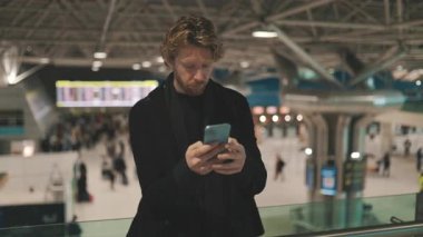 Endişeli genç işadamı cep telefonunu kontrol ediyor. Bu hareketli seyahat merkezinde hayal kırıklığını gösteriyor.