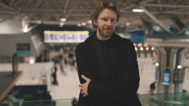 Uluslararası bir havaalanında duran ve kameraya bakan kızıl saçlı yakışıklı bir iş adamı.
