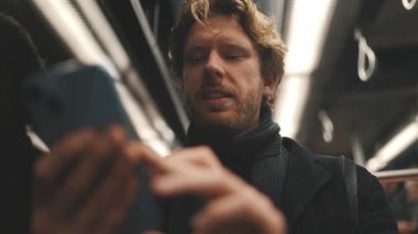 Toplu taşıma araçlarına gidip gelen kıvırcık saçlı ve gece akıllı telefonunu kullanan odaklanmış genç bir adam.