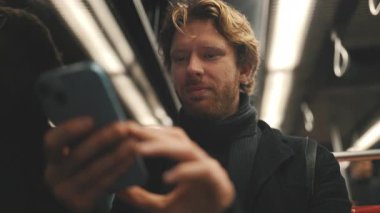 Geceleri metroda veya otobüste giderken cep telefonunu kullanan, sarı saçlı, gülümseyen bir adam.