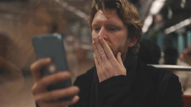 Telefonunu kullanan duygusal bir adam, metroda giderken şaşkınlık ve endişe gösteriyor.