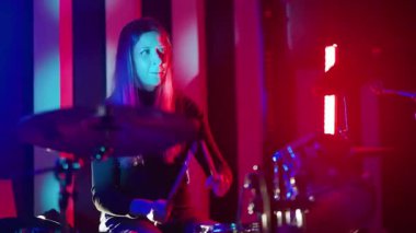 Karanlık bir stüdyoda davul çalan genç bir kadın kırmızı ve mavi neon ışıklarla canlı bir hava yaratıyor.