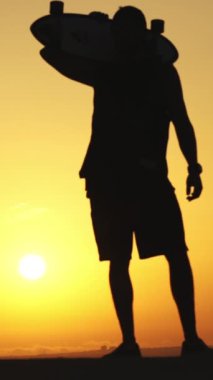 Omzunda kaykayıyla gün batımında duran genç bir adamın silueti. Orta çekim