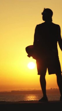 Gün batımında elinde kaykayla duran genç bir adamın silueti. Orta çekim