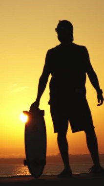 Parlak gün batımında elinde kaykayla duran genç bir atletin silueti. Orta çekim