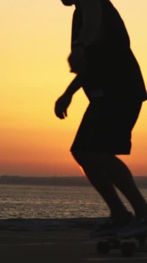 Okyanusun kenarında, parlak turuncu günbatımında uzun bir kaykayın üzerinde kayan bir adamın silueti. Orta çekim