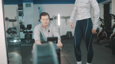 Yaşlı kadın spor salonunda kürek çekerken koçundan talimat alıyor.