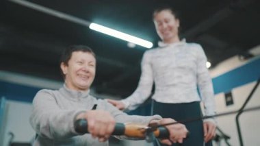 Gülümseyen yaşlı kadın, kadın spor hocasıyla birlikte kablolu yayında egzersiz yapıyor.