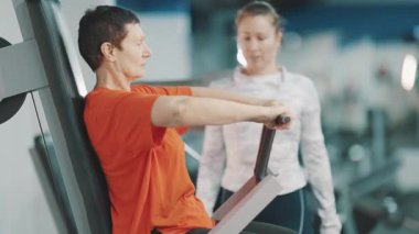 Yaşlı kadın spor salonunda kadın kişisel antrenörüyle bir makinede göğüs egzersizi yapıyor.