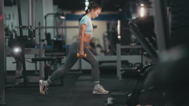 Modern bir fitness kulübünde spor yaparken dambıllarla hamle yapan kararlı genç bir kadın.