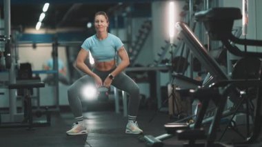 Modern bir fitness merkezinde kaslı bir kadın bir su ısıtıcısıyla güç antrenmanı yapıyor.