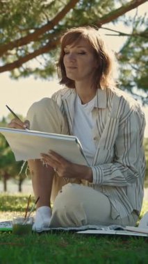 Yeşil parkta oturan ve çizim defterine resim çizen yetişkin bir kadın. Orta çekim