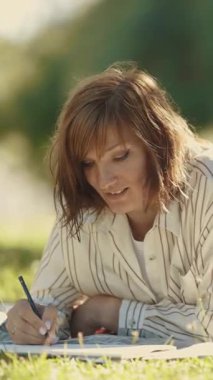 Bir kadın sanatçı çimlerin üzerinde uzanıyor, çizim defterine çiziyor ve gülümsüyor.