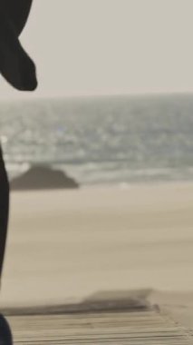 Dalgıç kıyafetli bir adam sahilde elinde sörf tahtasıyla yürüyor. Orta çekim