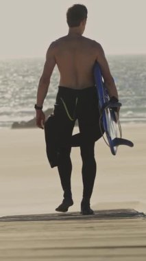 Dalgıç kıyafetli bir adam gün batımında elinde sörf tahtasıyla sahilde yürüyor. Orta çekim