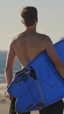 Sahilde çıplak gövdeli bir adam elinde sörf tahtasıyla dikiliyor. Orta çekim