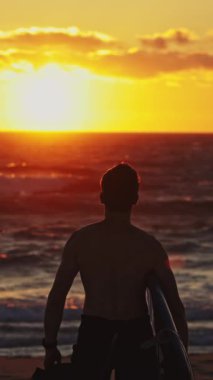 Parlak turuncu gün batımında okyanusa doğru yürüyen bir sörfçünün silüeti. Orta çekim