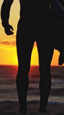 Parlak turuncu gün batımında okyanusa doğru yürüyen bir sörfçünün silüeti. Orta çekim