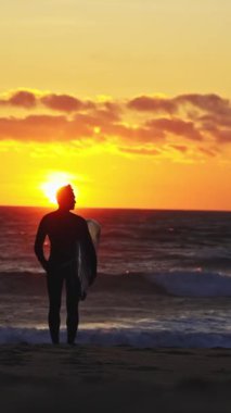 Turuncu gün batımında okyanusta yürüyen bir sörfçünün silüeti. Orta çekim