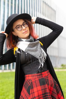 Dışarıda poz veren genç bir kadın, şapka, gözlük, eşarp ve etek giyiyor.