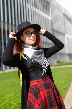 Bir kadın şapkasını düzeltiyor, kameraya bakıyor ve dışarıda gülümsüyor.