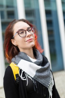 Kendine güvenen genç bir kadın yukarı bakıyor, gözlük takıyor ve açık havada eşarp takıyor.