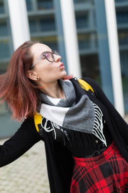 Genç bir kadın somurtuyor, gözlük takıyor, eşarp takıyor ve kırmızı ekose etek giyiyor.