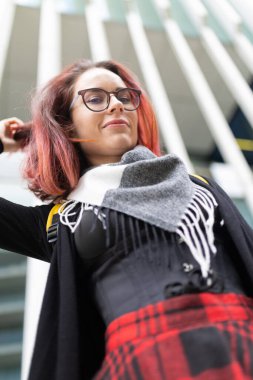 Kızıl saçlı ve gözlüklü genç bir kadın modern mimari arka plana bakıyor.