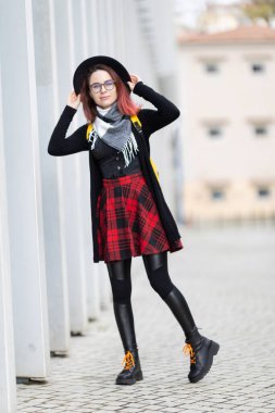 Şehir sokağında duran genç bir kadın, şapka, gözlük ve renkli kıyafetler giyiyor.